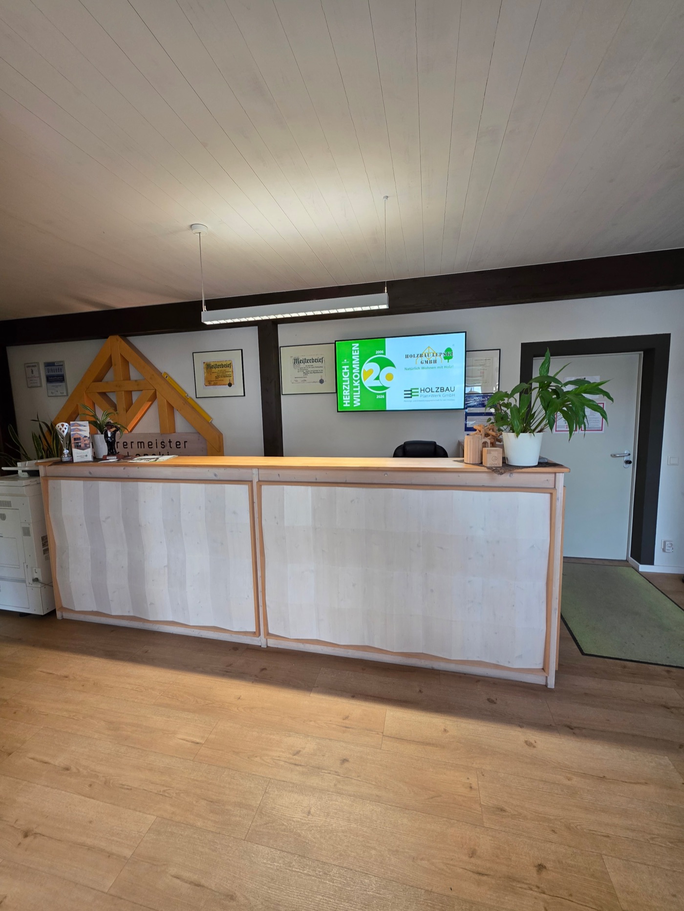 Empfangstresen aus hellem Holz in unserem Büro, dahinter ein Bildschirm mit den Logos der Holzbau Lepski GmbH und der Holzbau Plan+Werk GmbH und gerahmte Meisterbriefe