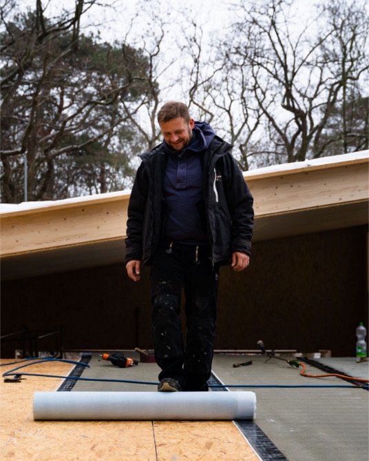 Dachdecker in Arbeitsjacke rollt auf einer Dachfläche aus Holzwerkstoffplatten eine Bahn aus