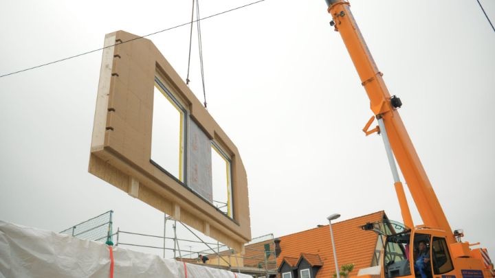 Vorgefertigtes Wandelement mit Fensteröffnung hängt am Kran über dem Rohbau eines Einfamilienhauses