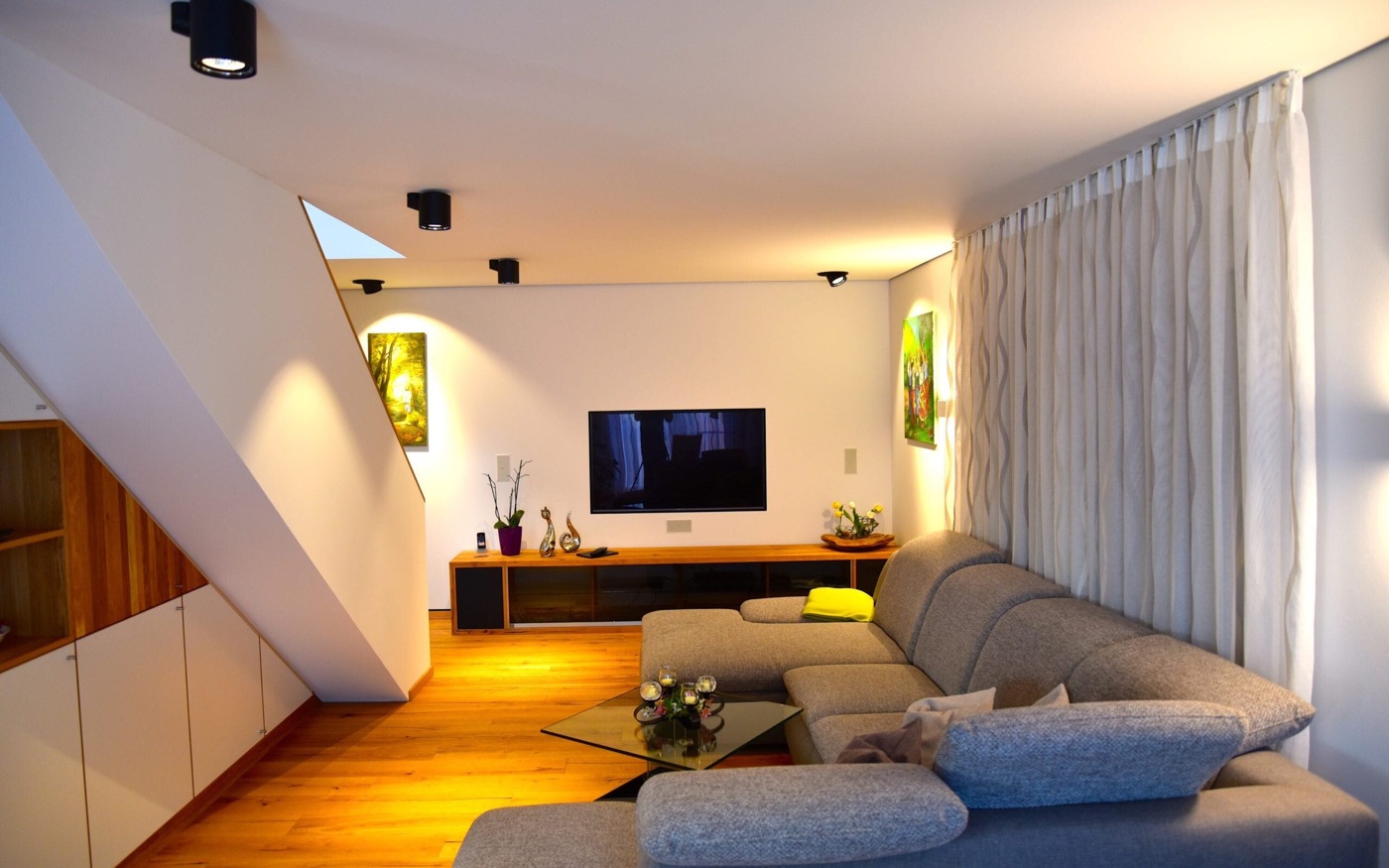 Wohnzimmer im Musterhaus „Felix“ am Abend: Ecksofa, Holzboden und Einbauschränke unter der Treppe